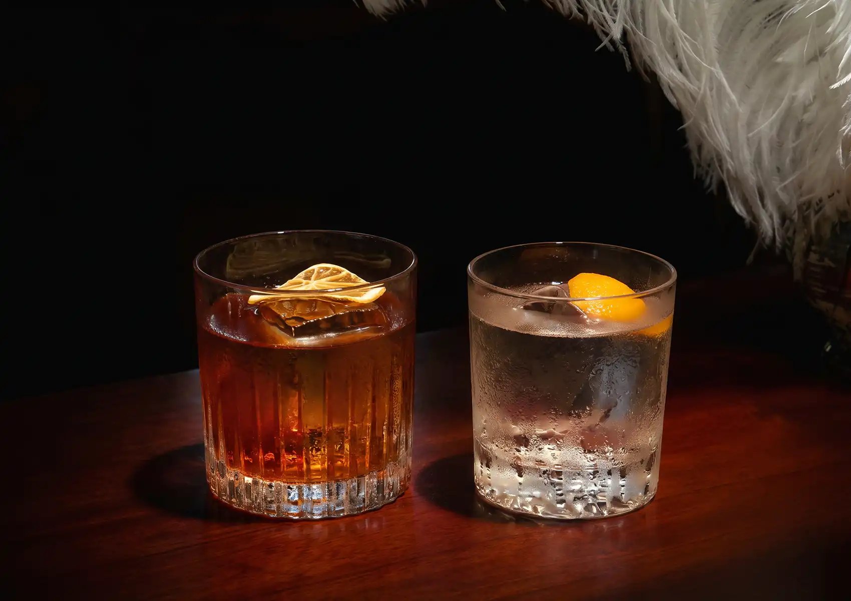 Two beautifully crafted drinks in short glass tumblers, one amber with a dehydrated citrus wheel and one clear with an orange peel twist, sitting on a dark wooden table at a Cocktail bar ubud.