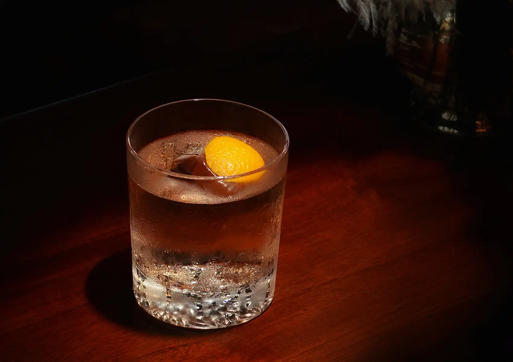 A clear cocktail over a large ice cube garnished with an orange peel in a rocks glass on a dark wooden surface at a cocktail bar ubud.