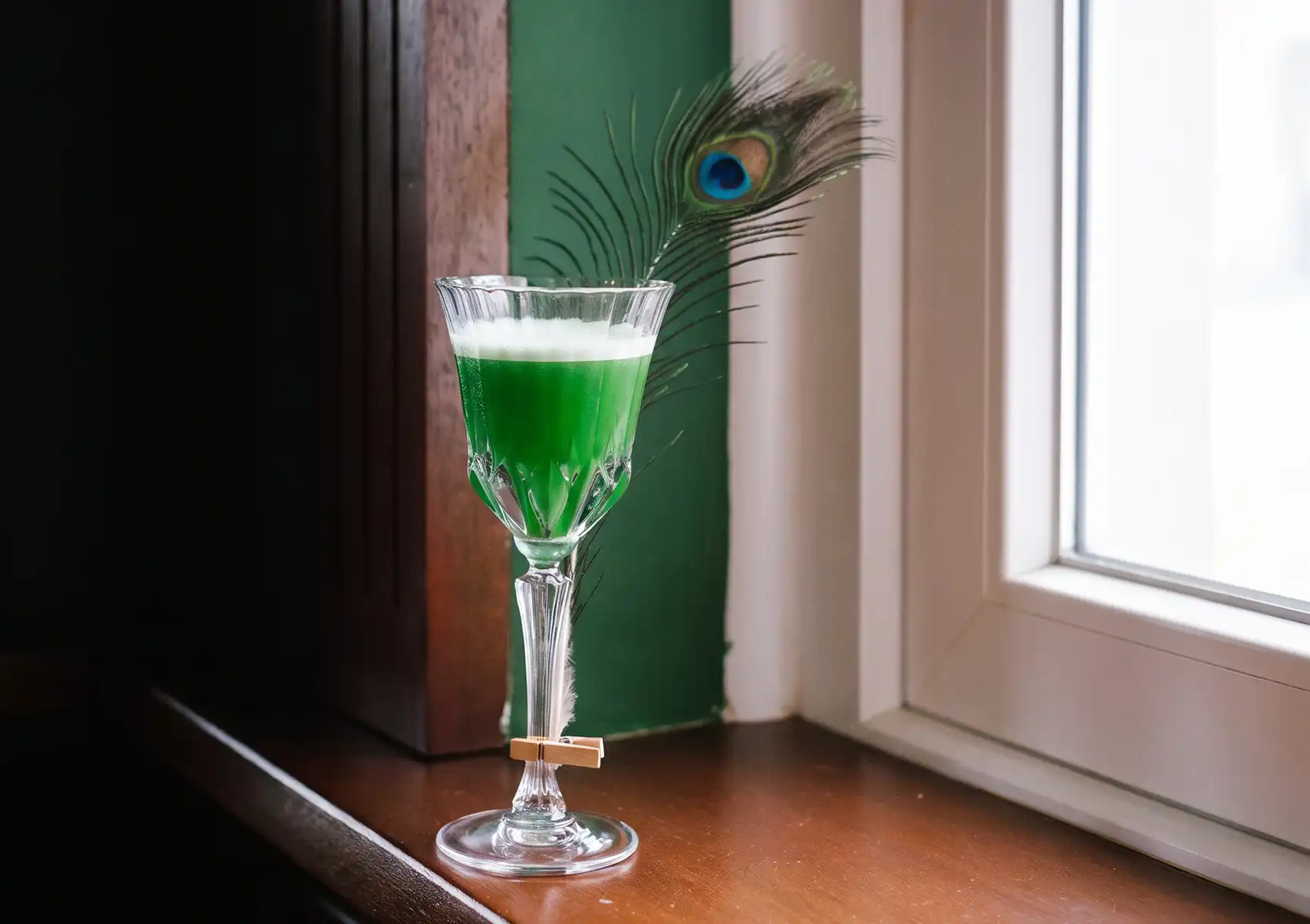 A bright green cocktail with a foamy top in an elegant stemmed glass garnished with a peacock feather, sitting on a wooden windowsill at a cocktail bar ubud.