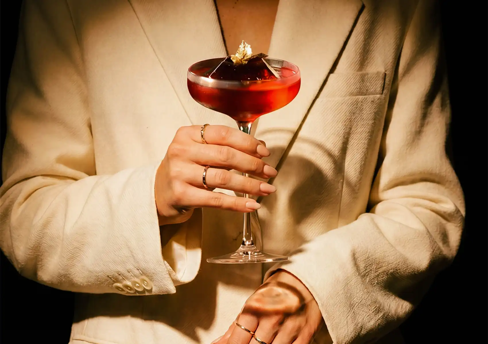 A person wearing a white suit jacket holding an elegant red cocktail topped with a large ice cube and gold leaf at a Cocktail bar ubud.