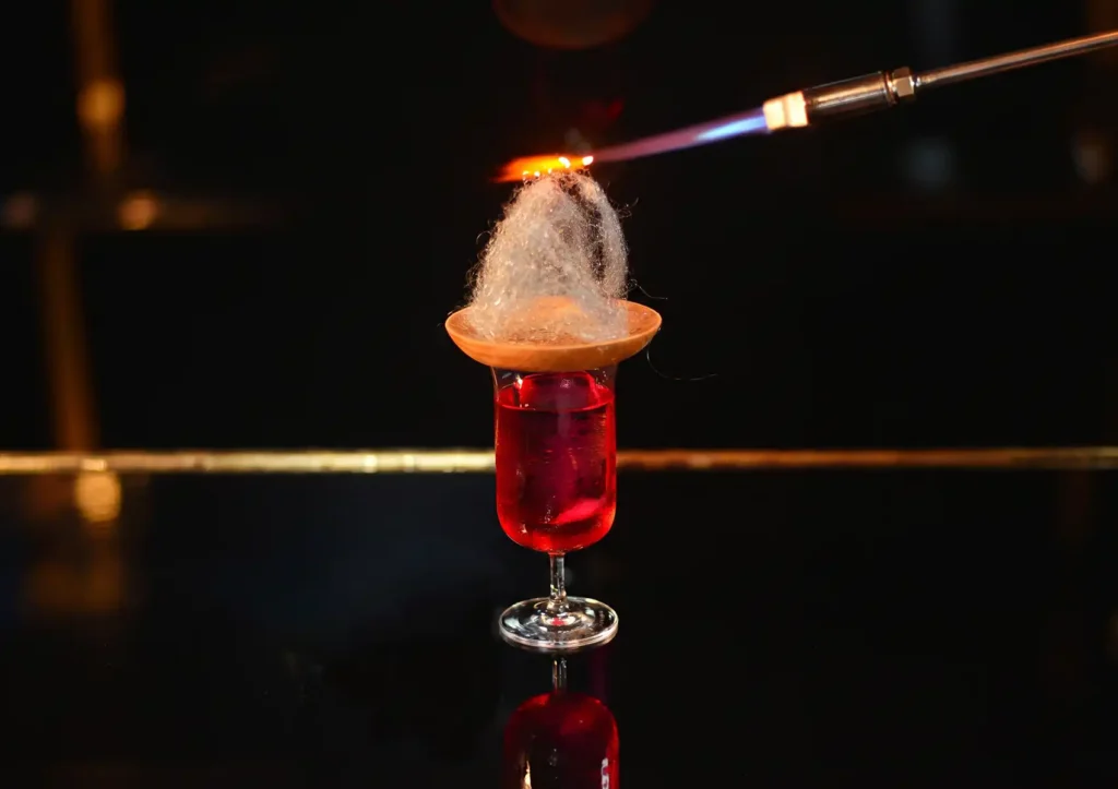 A vibrant red cocktail in a stemmed glass topped with a wooden disc holding spun sugar that is being carefully toasted by a blowtorch flame at a Cocktail bar ubud.