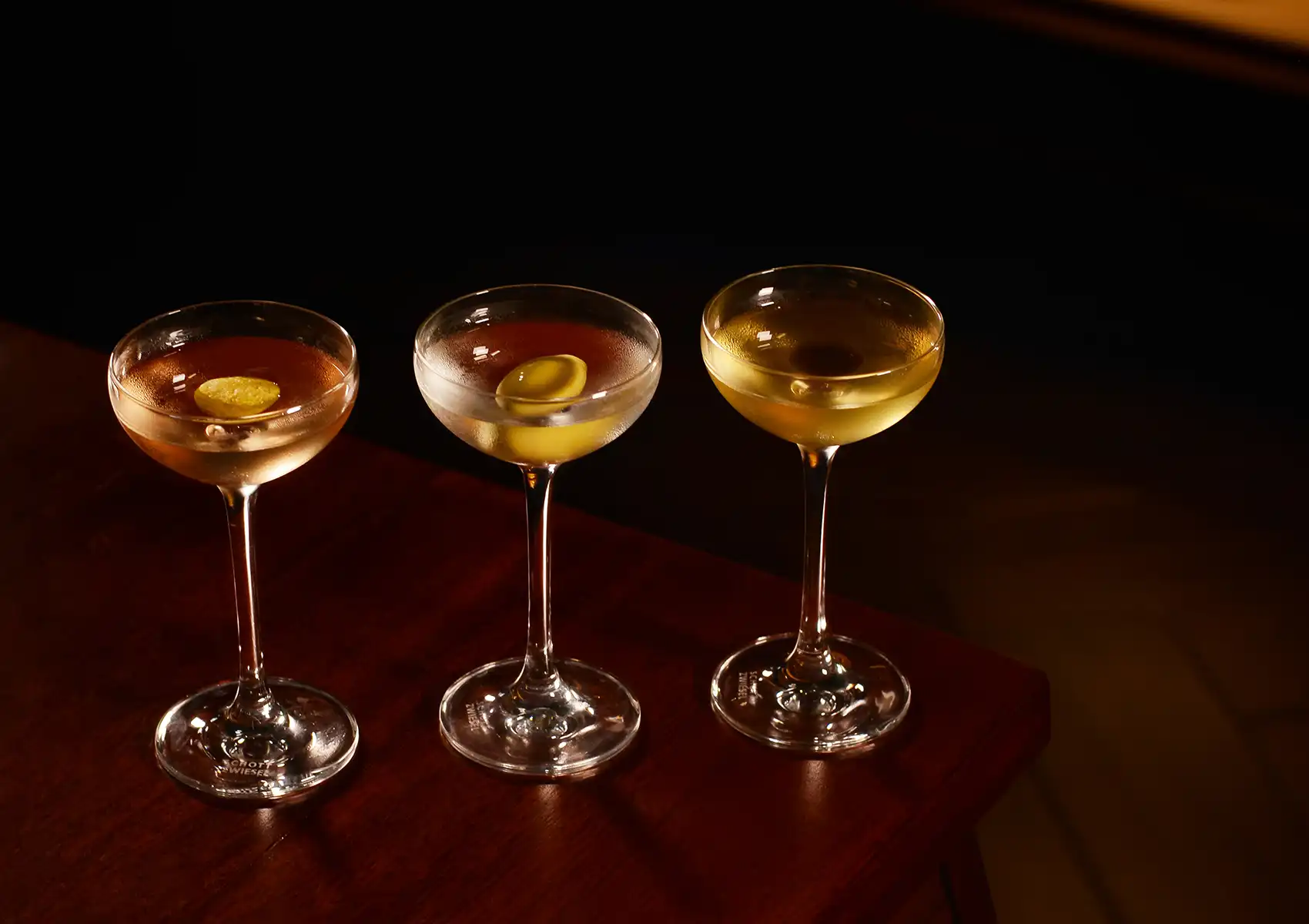 Three elegant clear cocktails in coupe glasses, featuring citrus and olive garnishes, lined up on a dark wooden table at a Cocktail bar ubud.