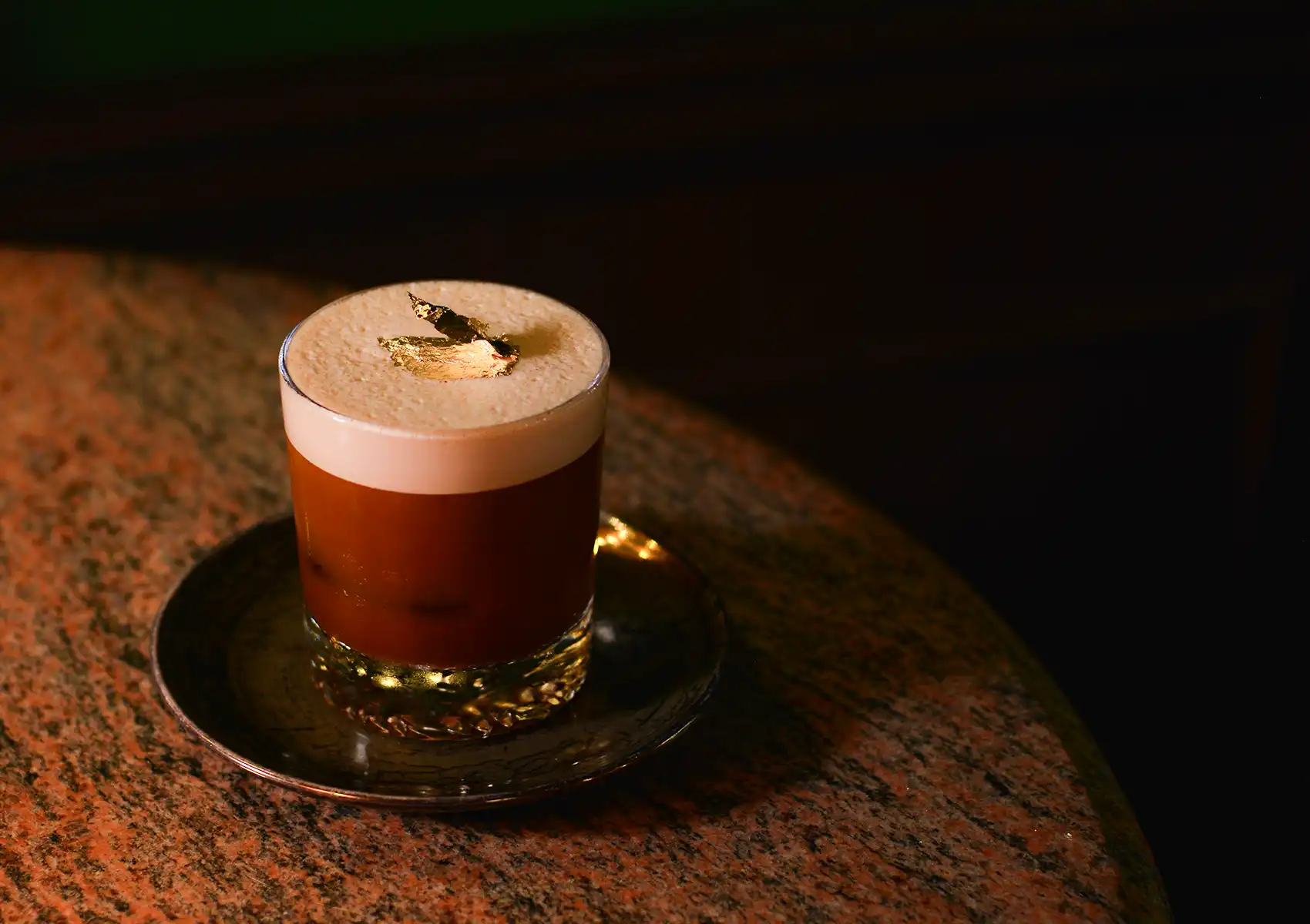 A close-up of a frothy cocktail topped with a gold leaf garnish on a textured table inside a dimly lit cocktail bar ubud.