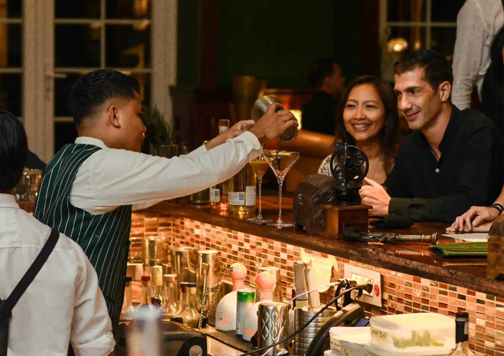 A smiling couple sitting at a counter watching a bartender in a green pinstripe vest expertly pour a freshly shaken drink into a martini glass at a lively Cocktail bar ubud.